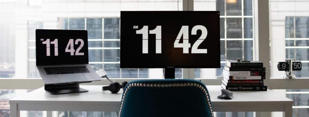 A photograph of a white desk in front of a large office window. A laptop and computer monitor displaying the time are on the desk.