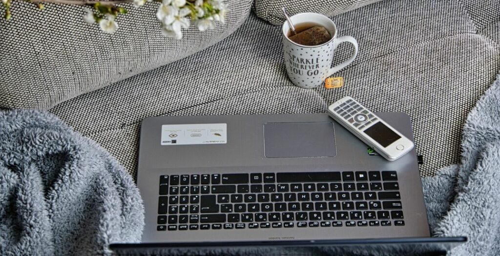 A photograph of a laptop, a cup of tea, a blanket, and an old cell phone on a couch.