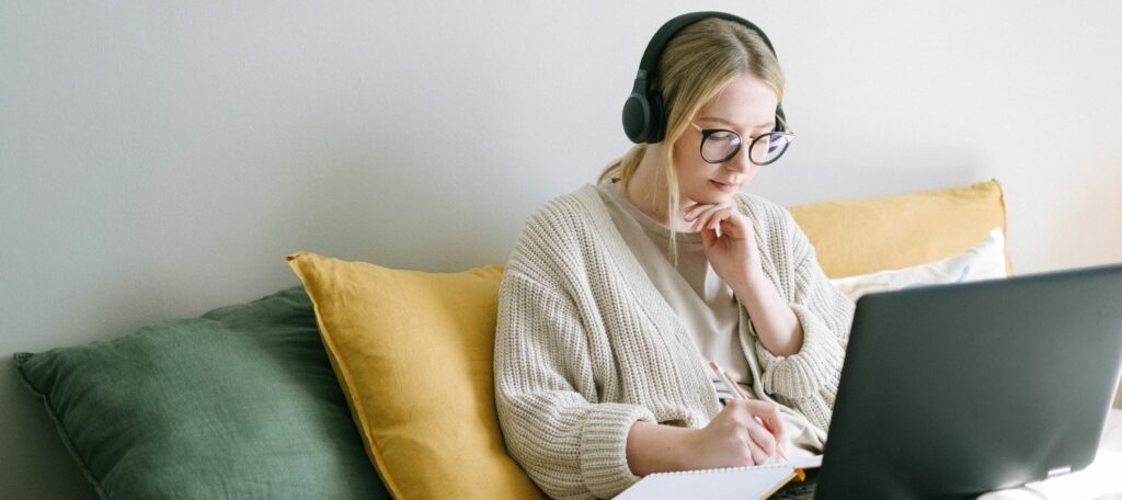 A young woman is sitting on her couch, wearing headphones, and working on her laptop.