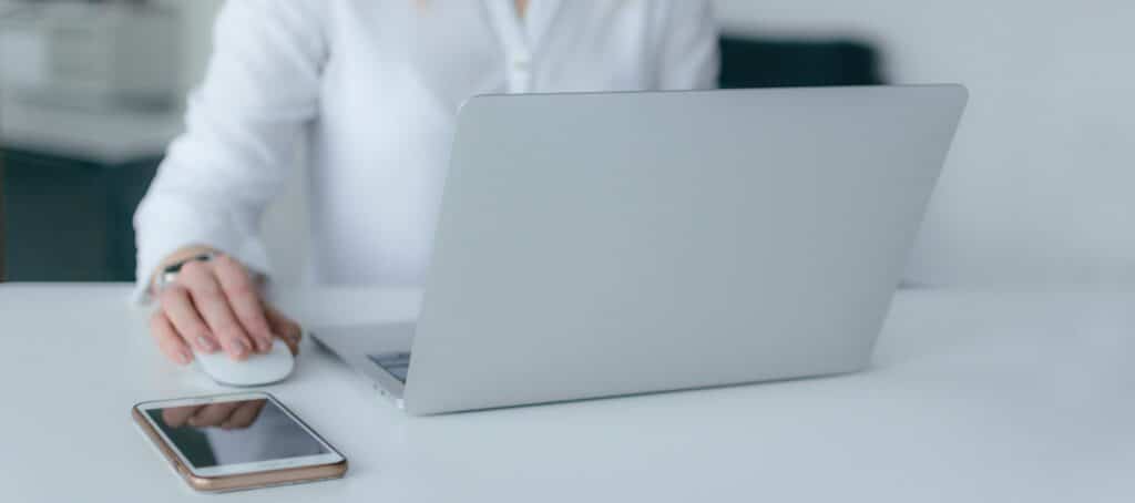 A photo of a woman in white working on her laptop beside her mobile phone.