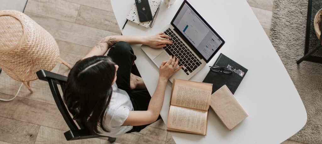 An overhead photo of a young woman working on her laptop at a desk with open books.