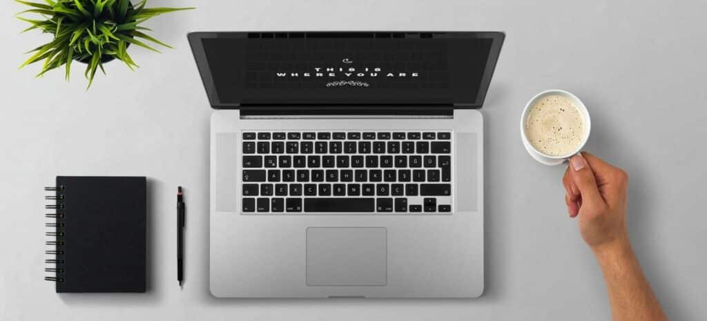 An overhead photograph of a notebook, laptop, and someone holding a cup of coffee at a white desk.