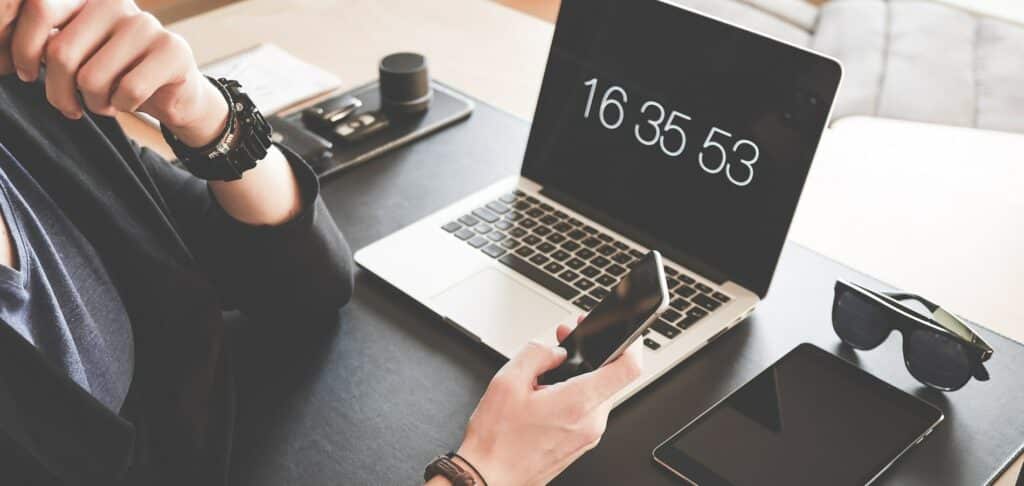 A photograph of someone looking at their phone while sitting at their desk with a laptop.