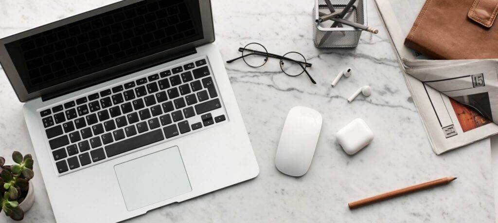 A overhead shot of a laptop, glasses, and a computer mouse and other random office accessories on a white, marble desk.