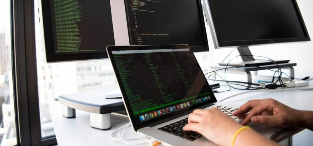 A photograph of someone typing on a laptop with large computer monitors in the background