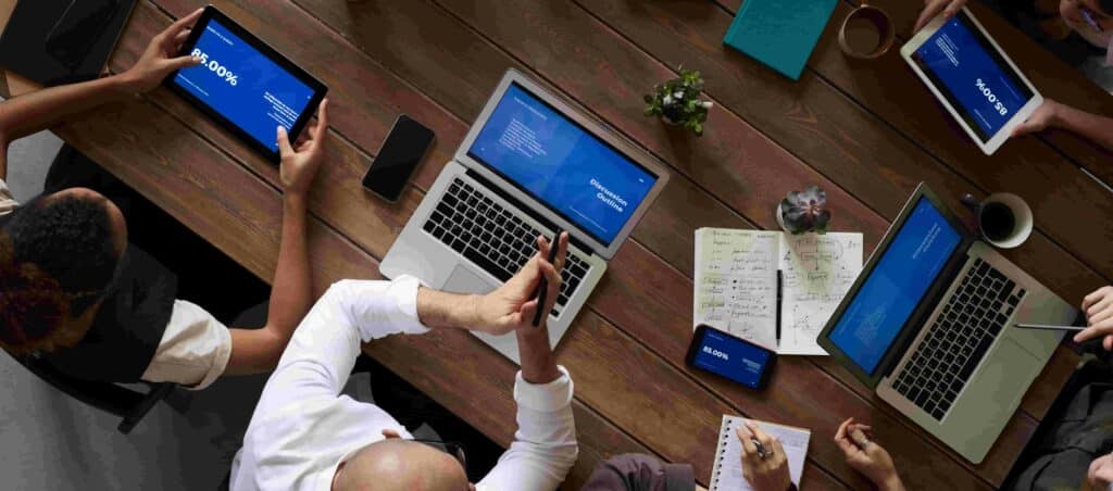 An overhead photograph of several people sitting at a large table working on laptops and tablets together.
