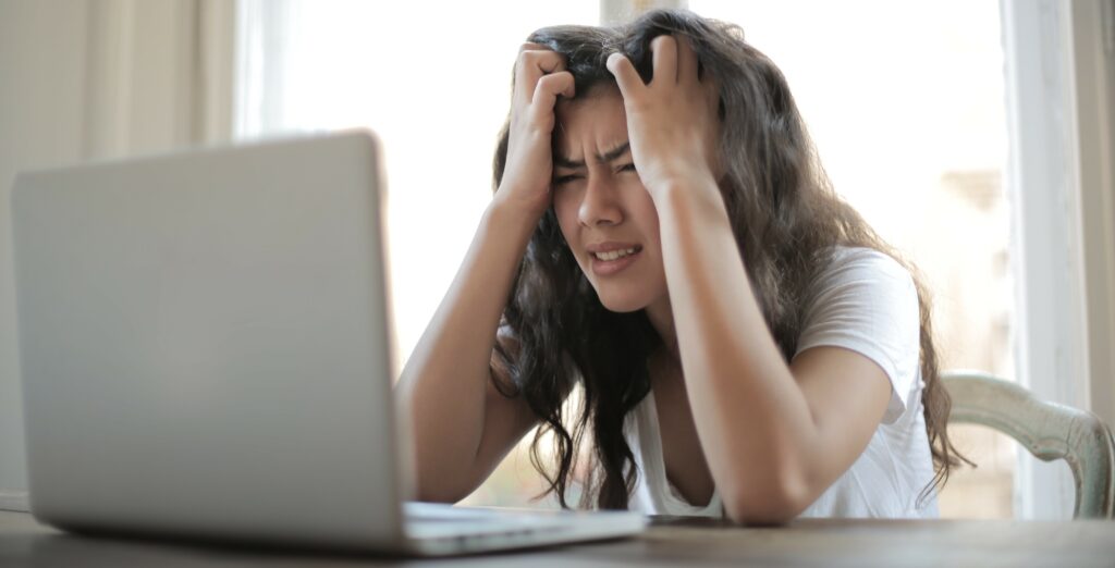 A woman holds her head in her hands and scowls at her laptop in frustration.