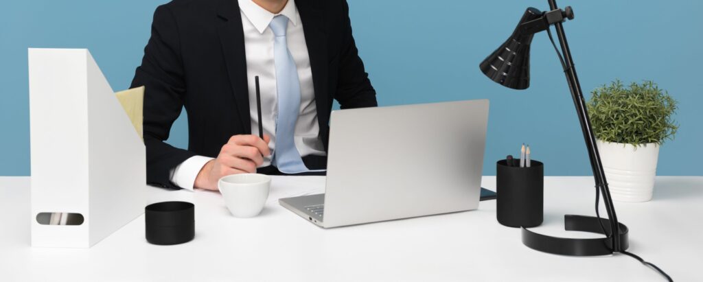 A photograph of a man in a suit sitting at a white desk working on a laptop.