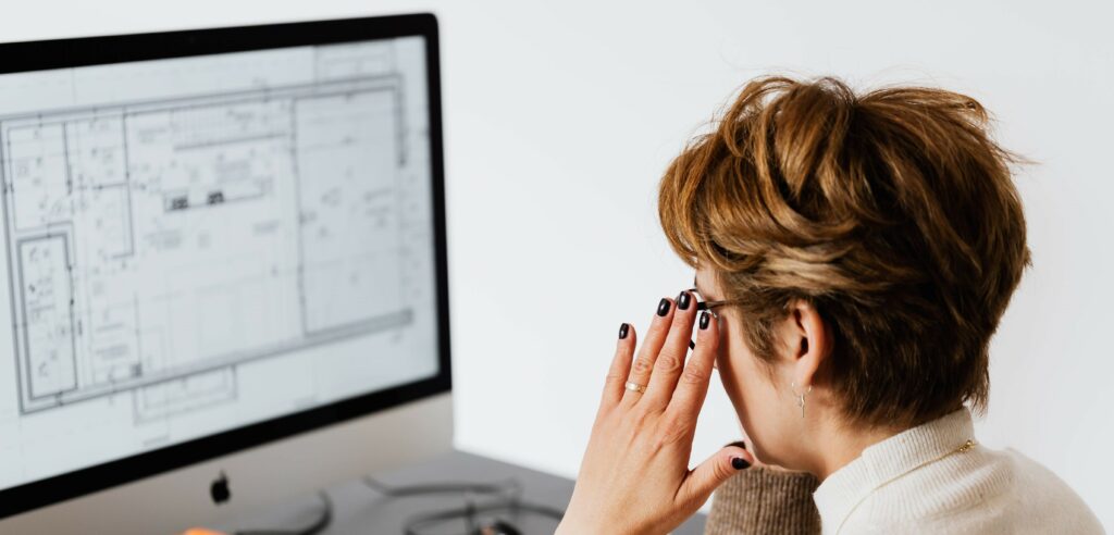 An over-the-shoulder photograph of a woman with short hair looking at a computer screen. Her hands are on her temples in thought or distress.