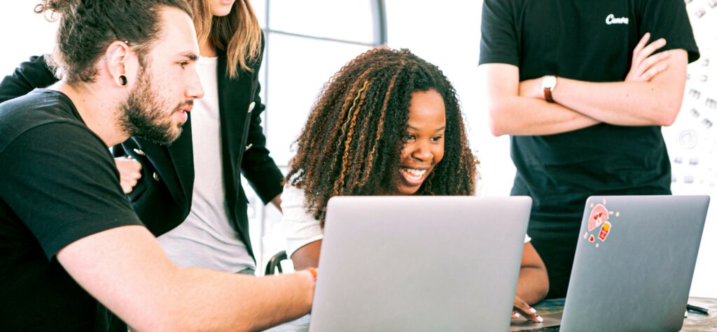 A photograph of a young woman smiling and working on her laptop surrounded by men and women watching her. 