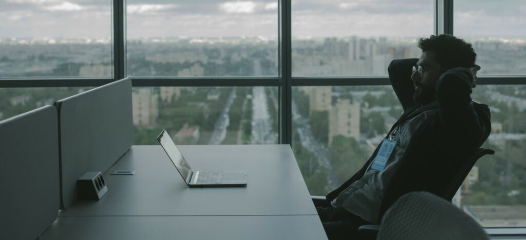 A man sits at an office cubicle looking at his laptop. He is seated next to a window overlooking a city.