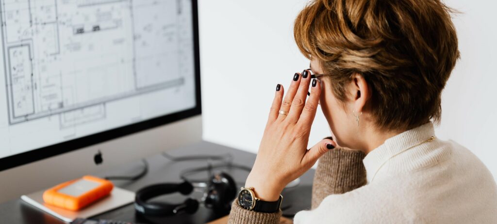 A photograph of a woman looking at her computer. She has her hands on her temples as if in shock or frustration.