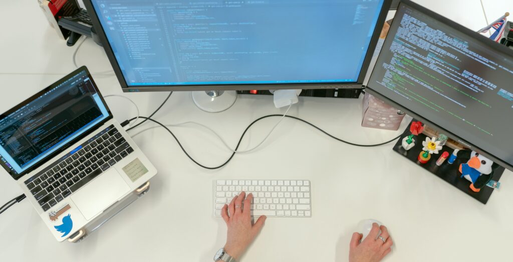 A woman sits at a white desk working on a laptop and multiple monitors