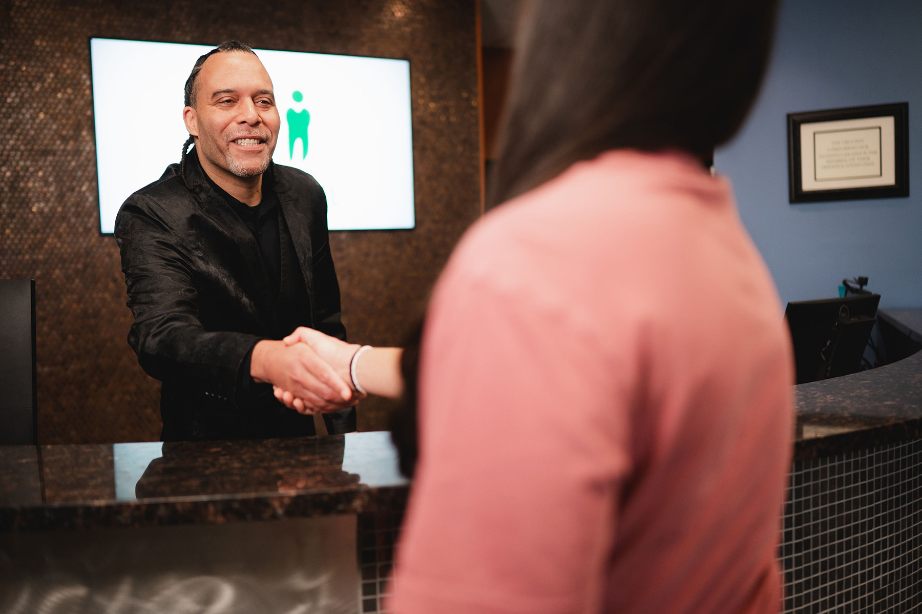 Smiling man with braided hair shaking hands with a woman across a reception desk in an office.