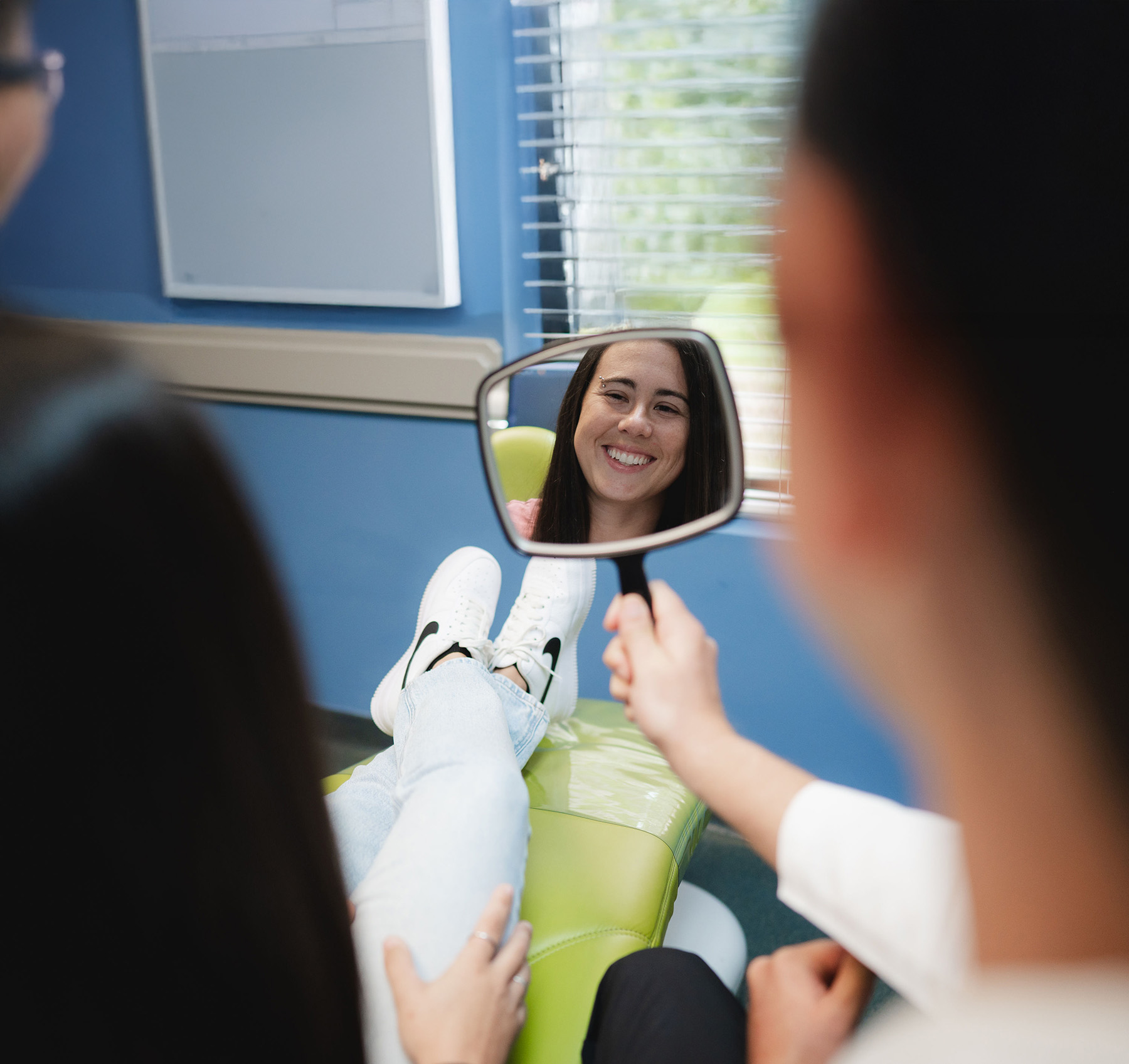 Smiling woman with dark hair reflected in a dental mirror held by a person in a dental office.