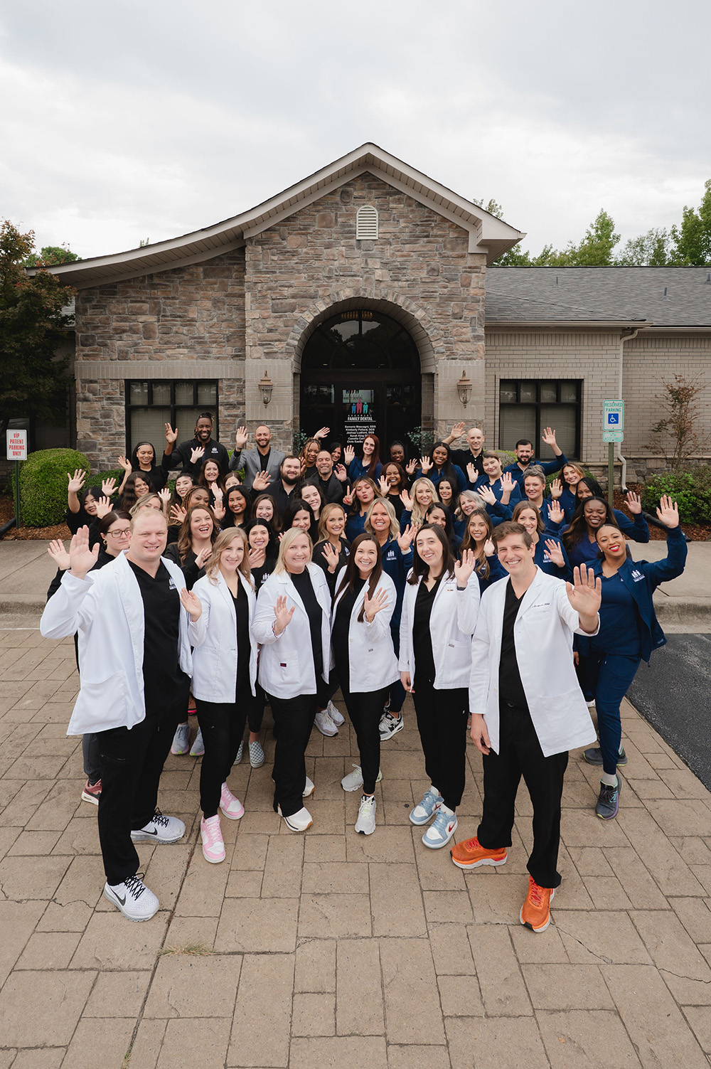 Large group of smiling dental professionals waving, standing outside a stone dental office building.