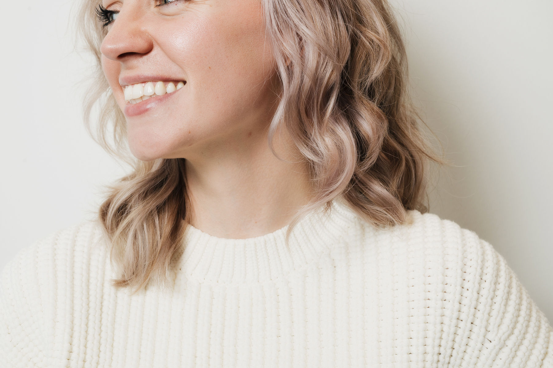 Close-up of a smiling woman with wavy blonde hair wearing a cream knit sweater, facing left.