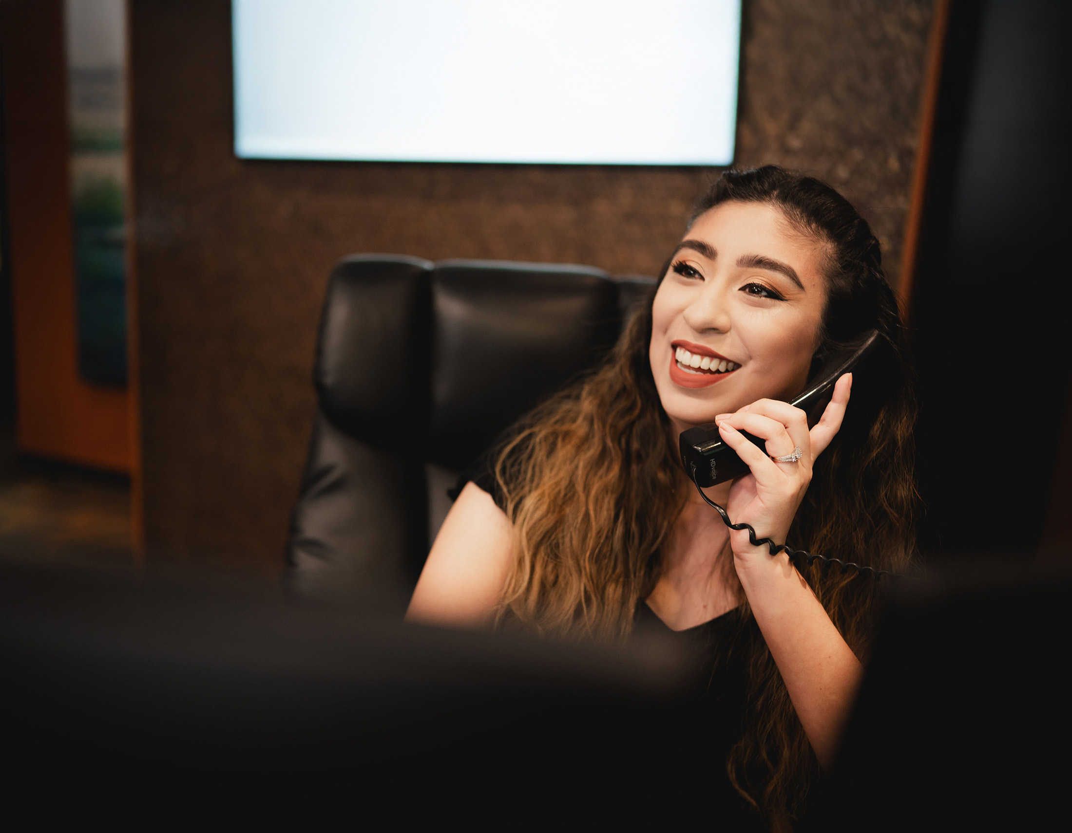 Smiling woman with long wavy hair talking on a corded phone in an office setting.