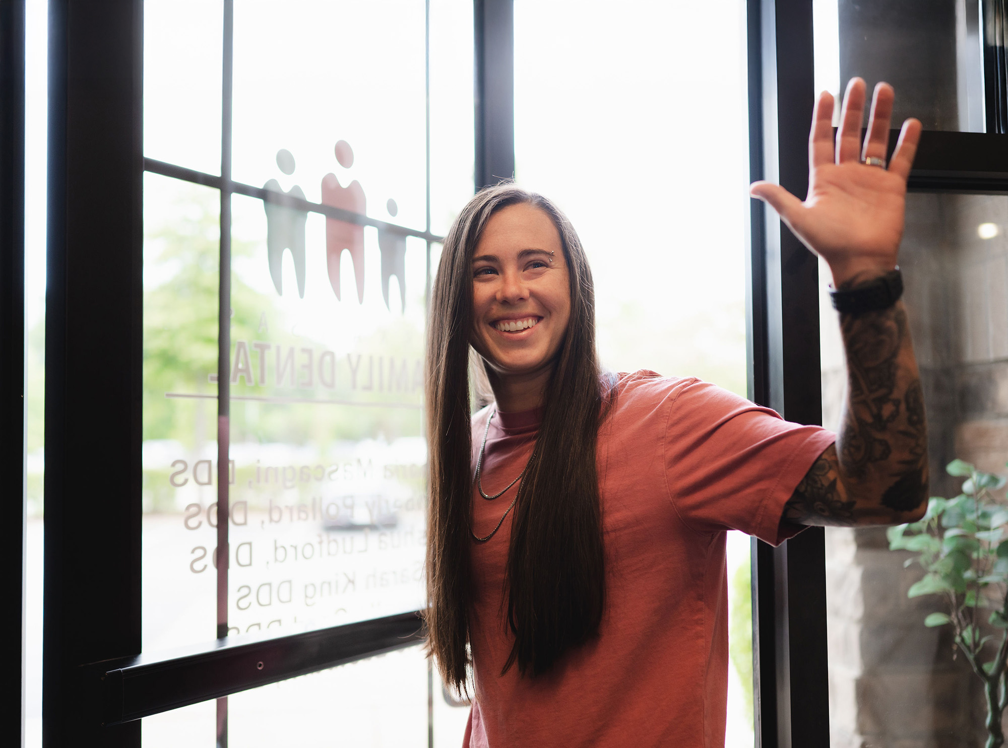 Smiling person with long hair and tattoos waving while standing near a glass door with dental office signage.