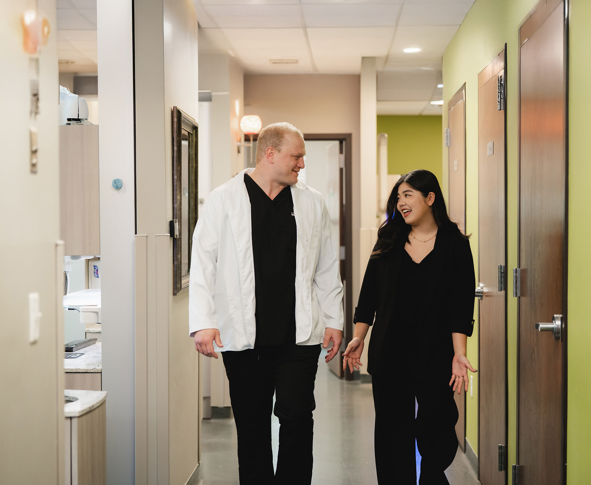 Smiling male dentist in a white coat walking and talking with a female colleague in black attire down a clinic hallway.