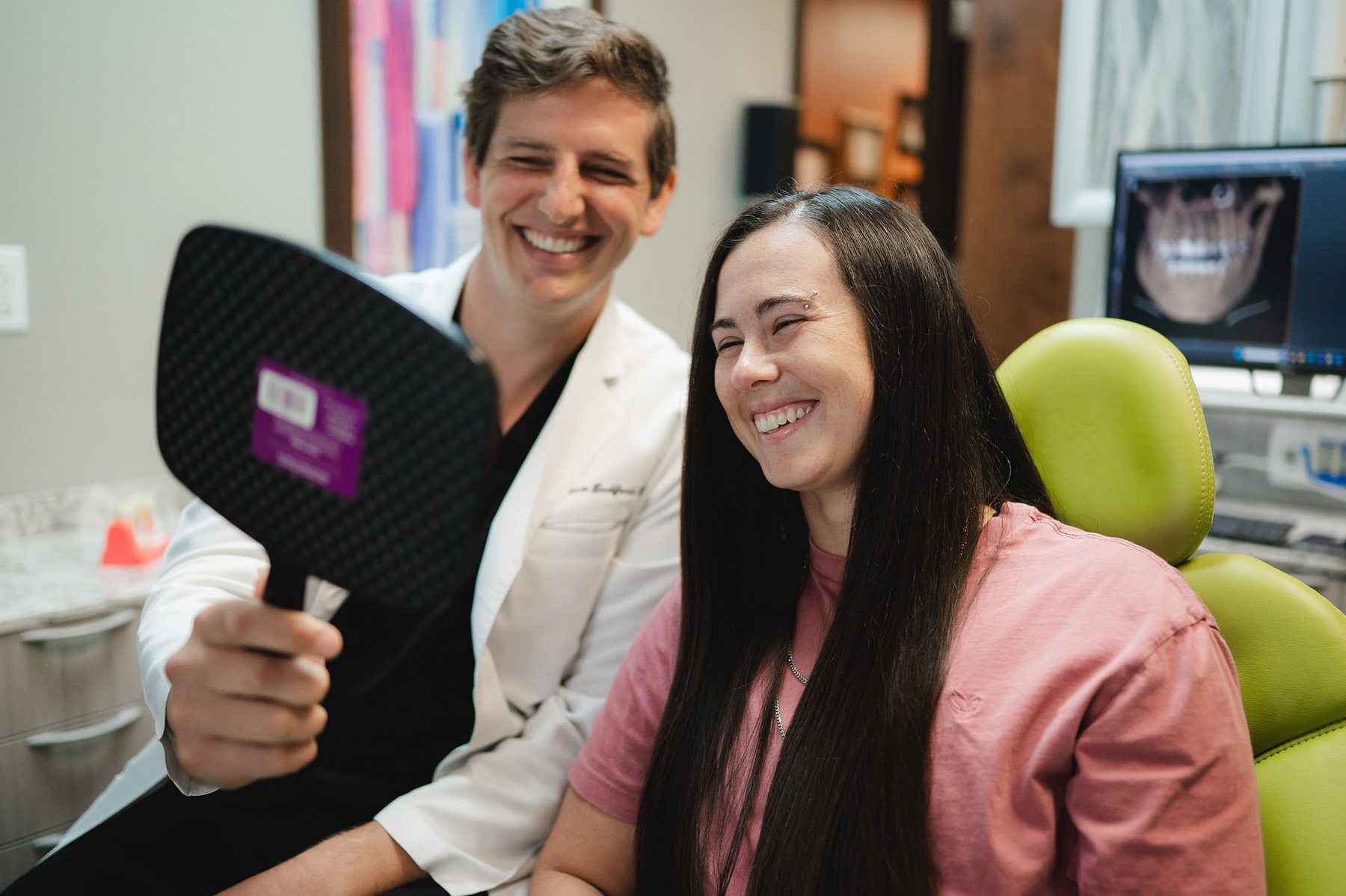 Dentist showing a hand mirror to a smiling female patient seated in a green dental chair.