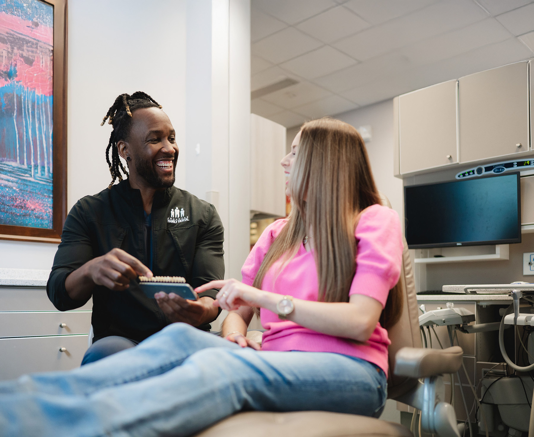 Dental professional showing a dental shade guide to a female patient seated in a dental chair, both smiling in a dental office.