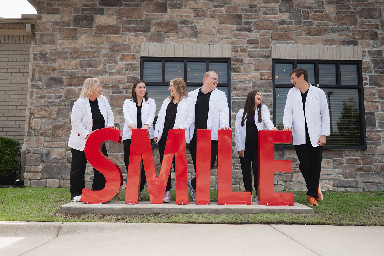 Six dental professionals in white coats standing behind large red letters spelling SMILE, smiling and interacting outside a stone building.