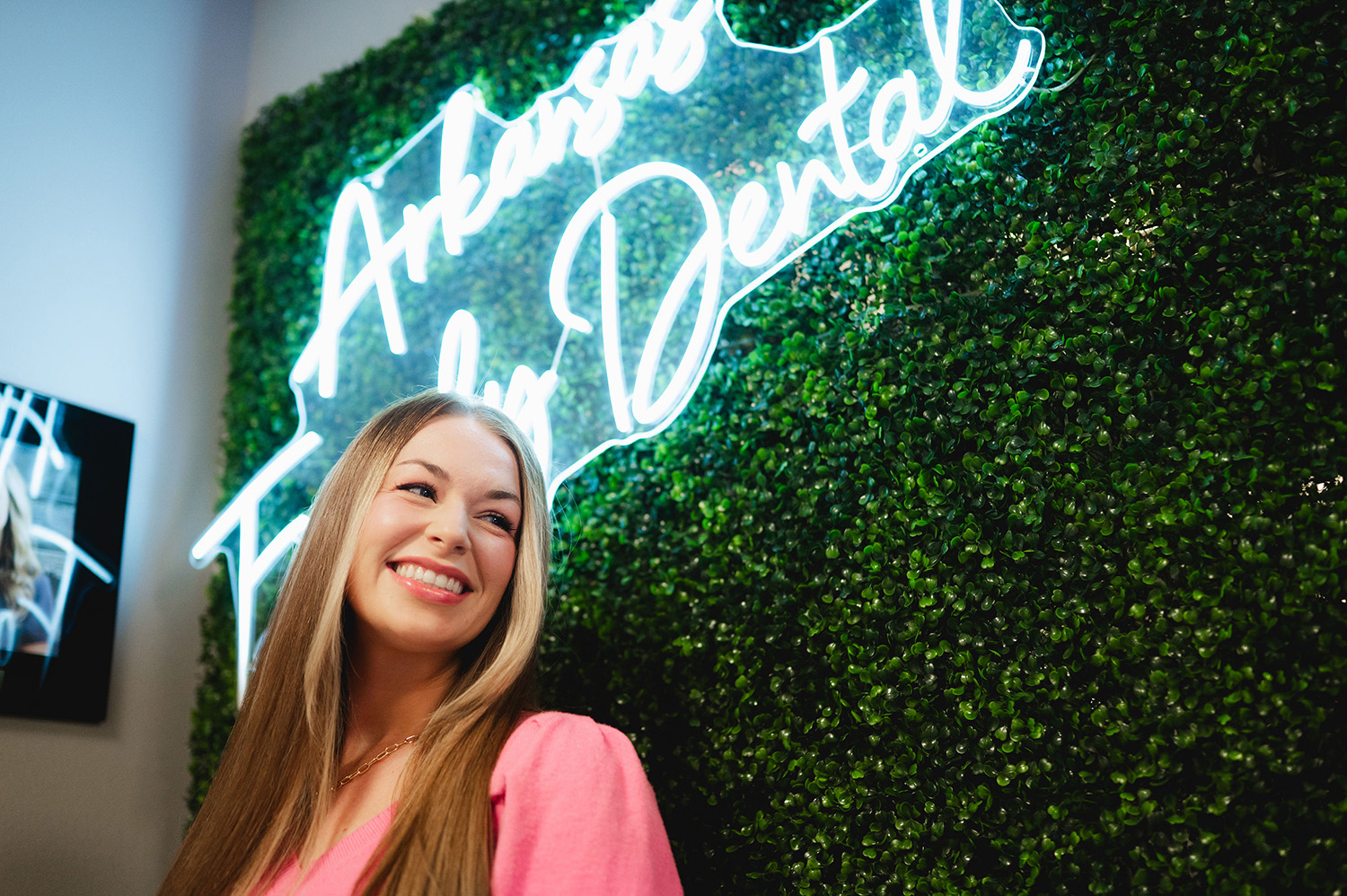 Smiling woman with long blonde hair standing in front of a green leafy wall with a glowing neon sign reading Arkansas Family Dental.