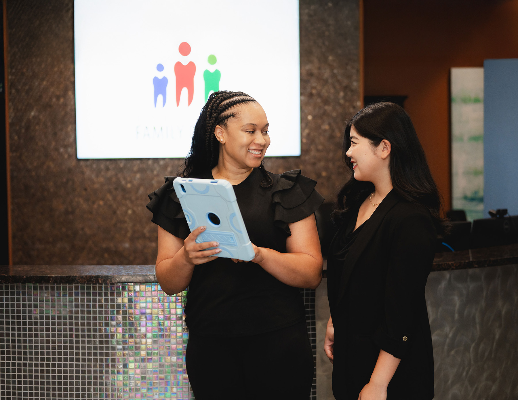 Two women smiling and talking in a professional setting, one holding a tablet, with a family dental logo in the background.