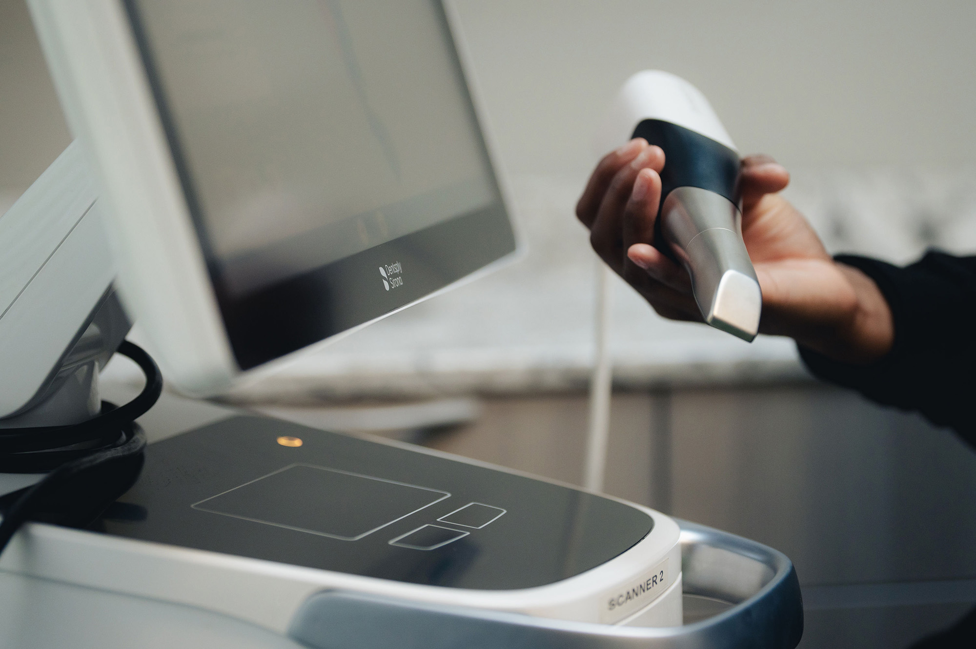 Hand holding a dental scanning device in front of a Dentsply Sirona monitor and scanner unit labeled SCANNER 2.