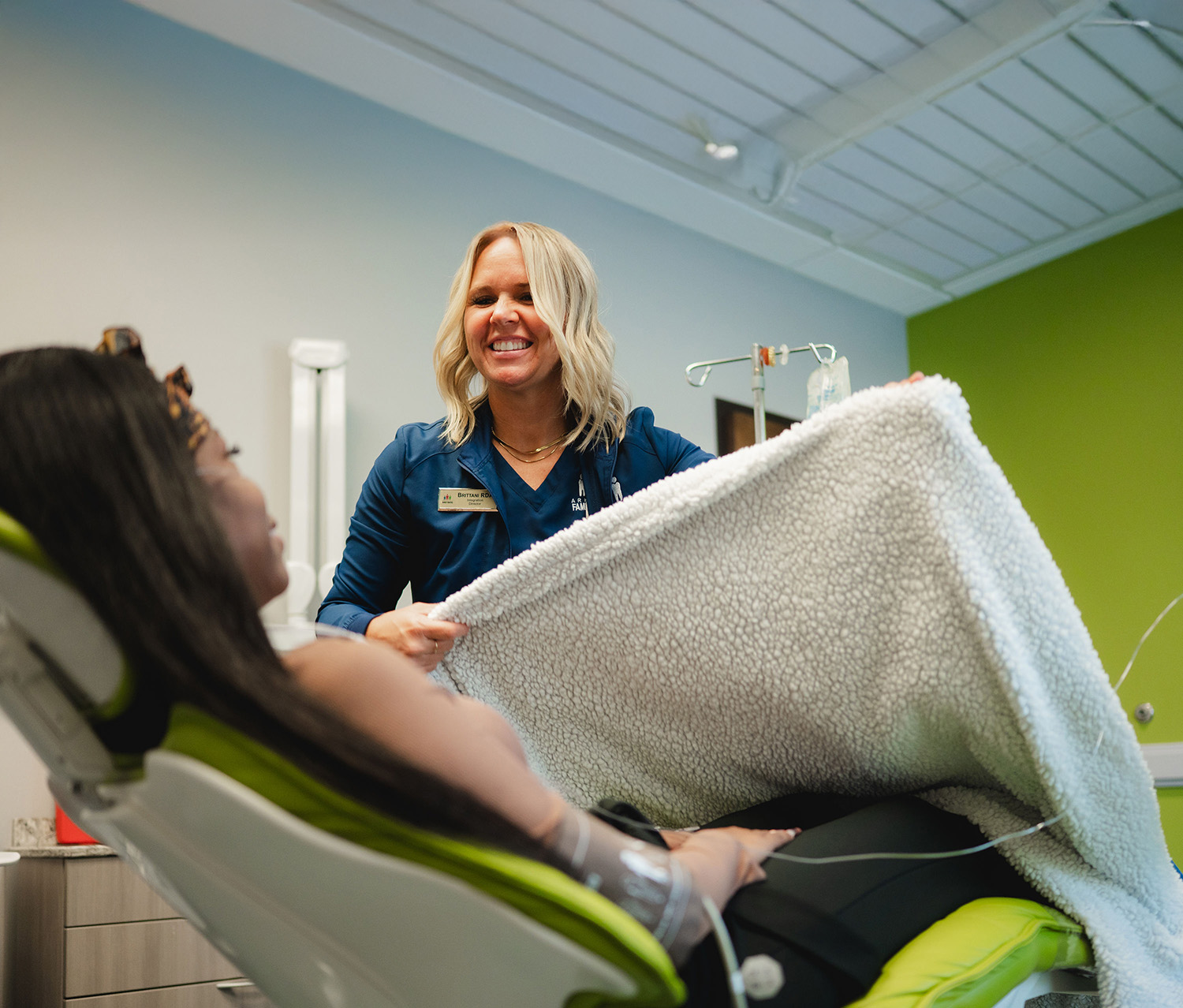 Smiling healthcare worker placing a blanket on a reclining patient with an IV drip in a medical room.