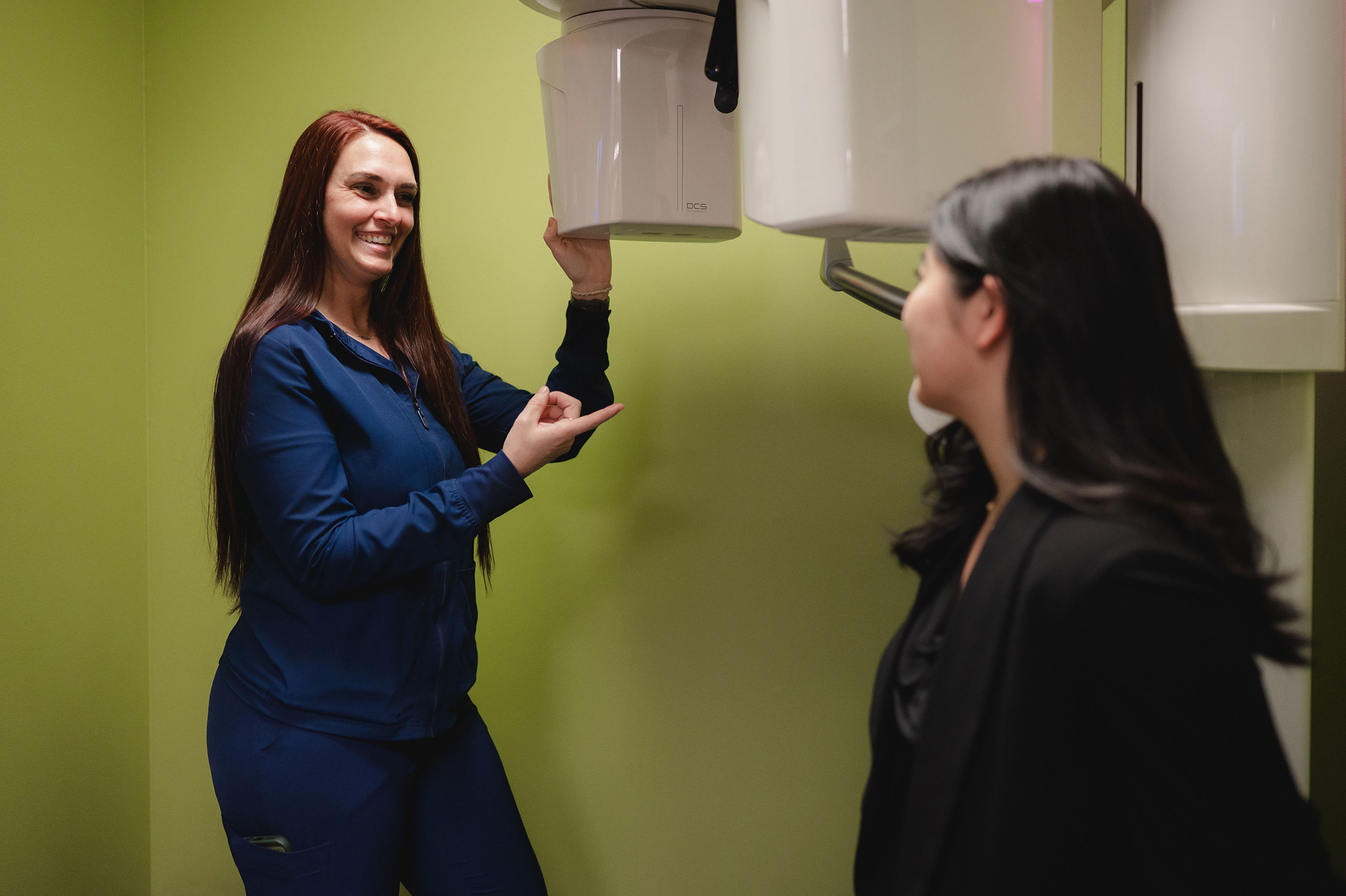 Smiling dental professional in blue scrubs adjusting dental X-ray machine while talking to a patient.