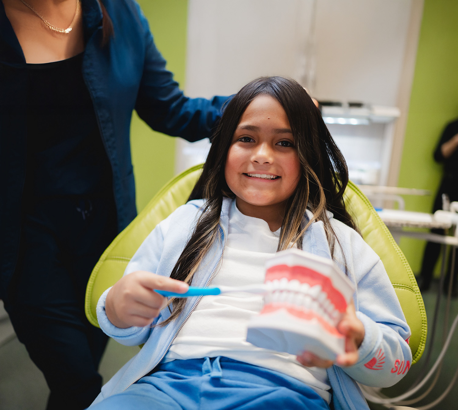 Smiling girl sitting in a dental chair holding a toothbrush and a dental model of teeth.