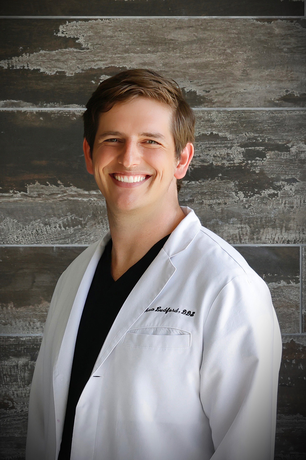 Smiling man wearing a white lab coat over black scrubs, standing against a textured dark wall.