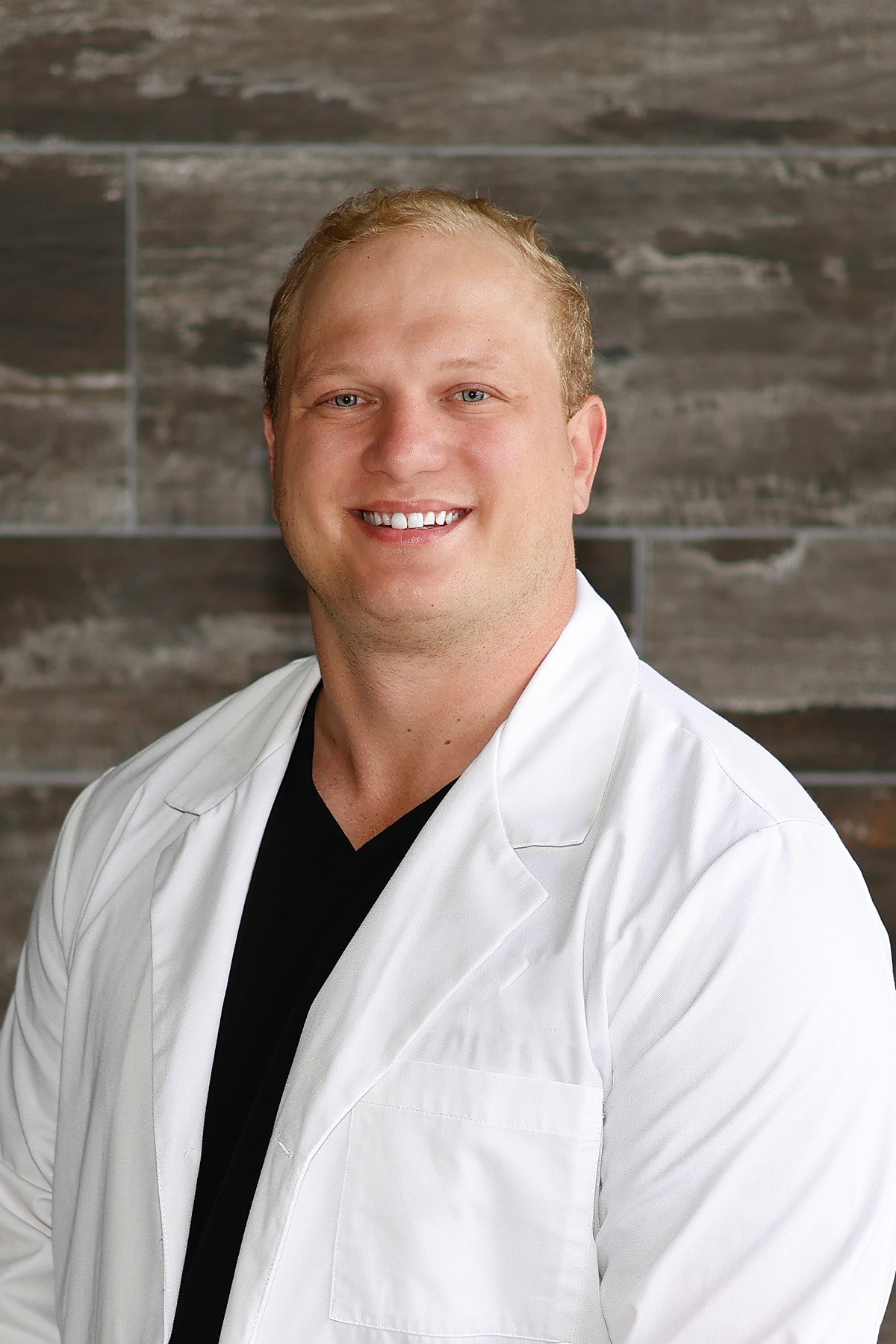 Smiling man with short blond hair wearing a white lab coat over a black shirt in front of a textured gray wall.