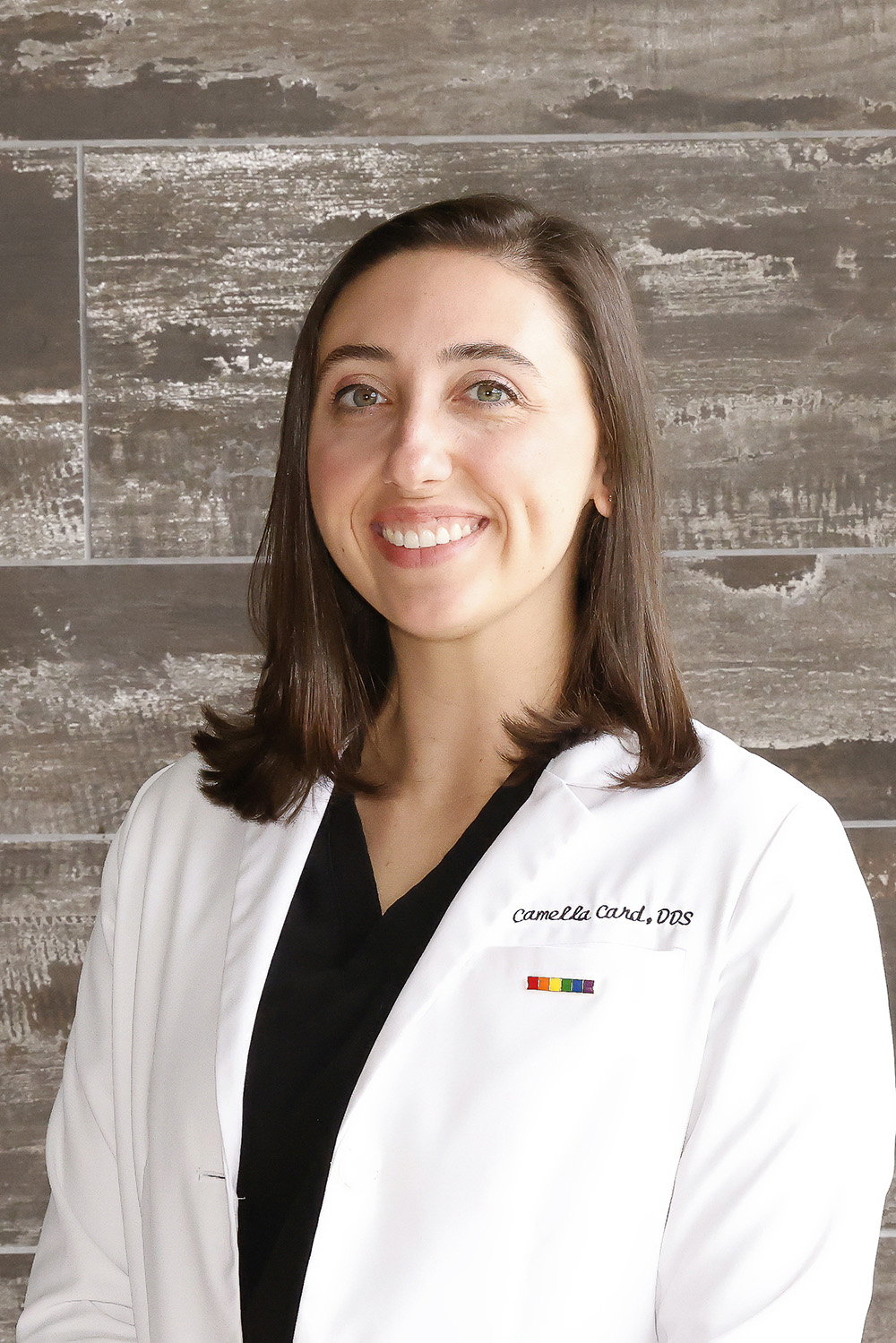 Smiling woman with brown hair wearing a white lab coat embroidered with 'Camella Card, DDS' and a rainbow pin.