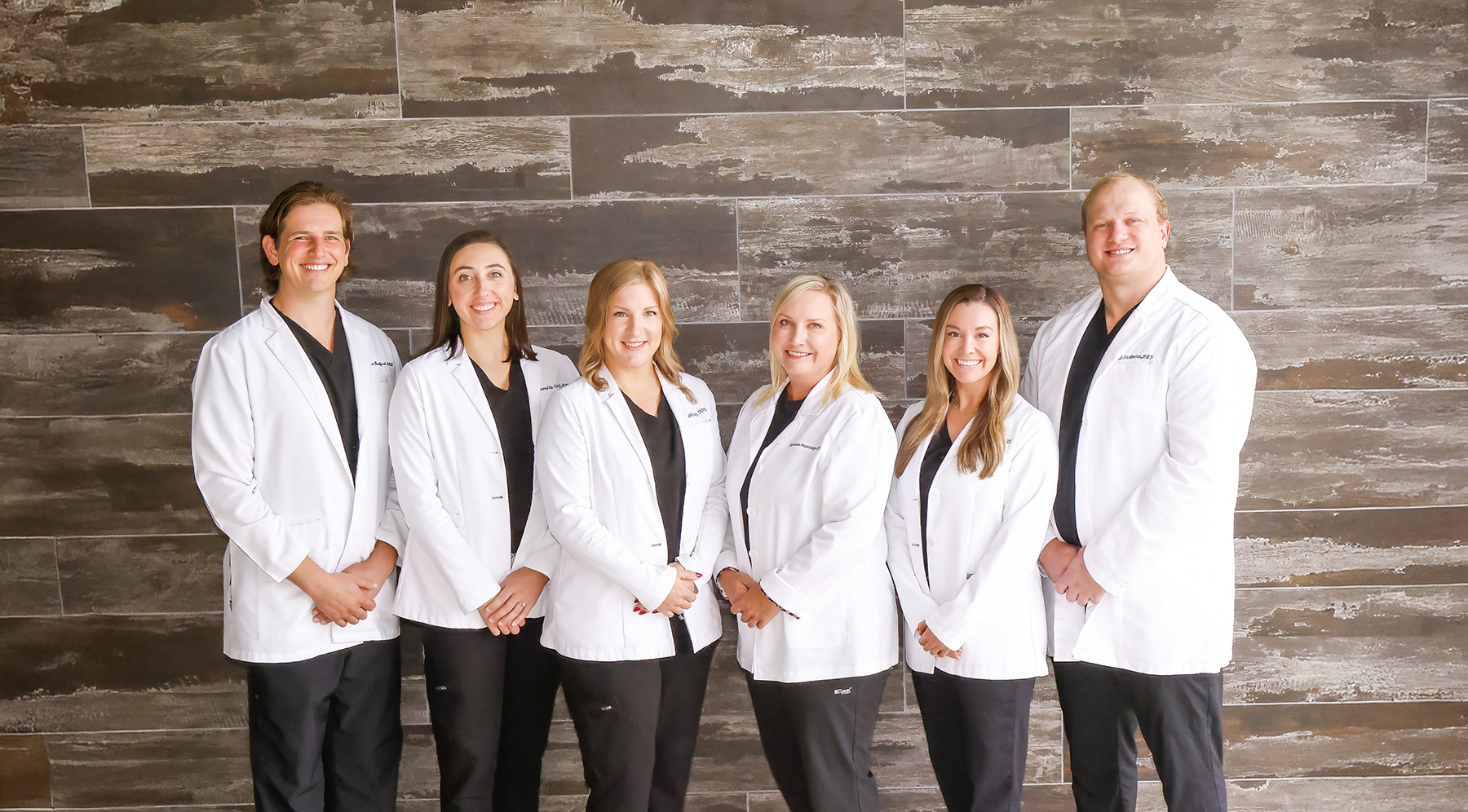 A team of six medical professionals standing and smiling, wearing white coats and black scrubs against a stone wall background.