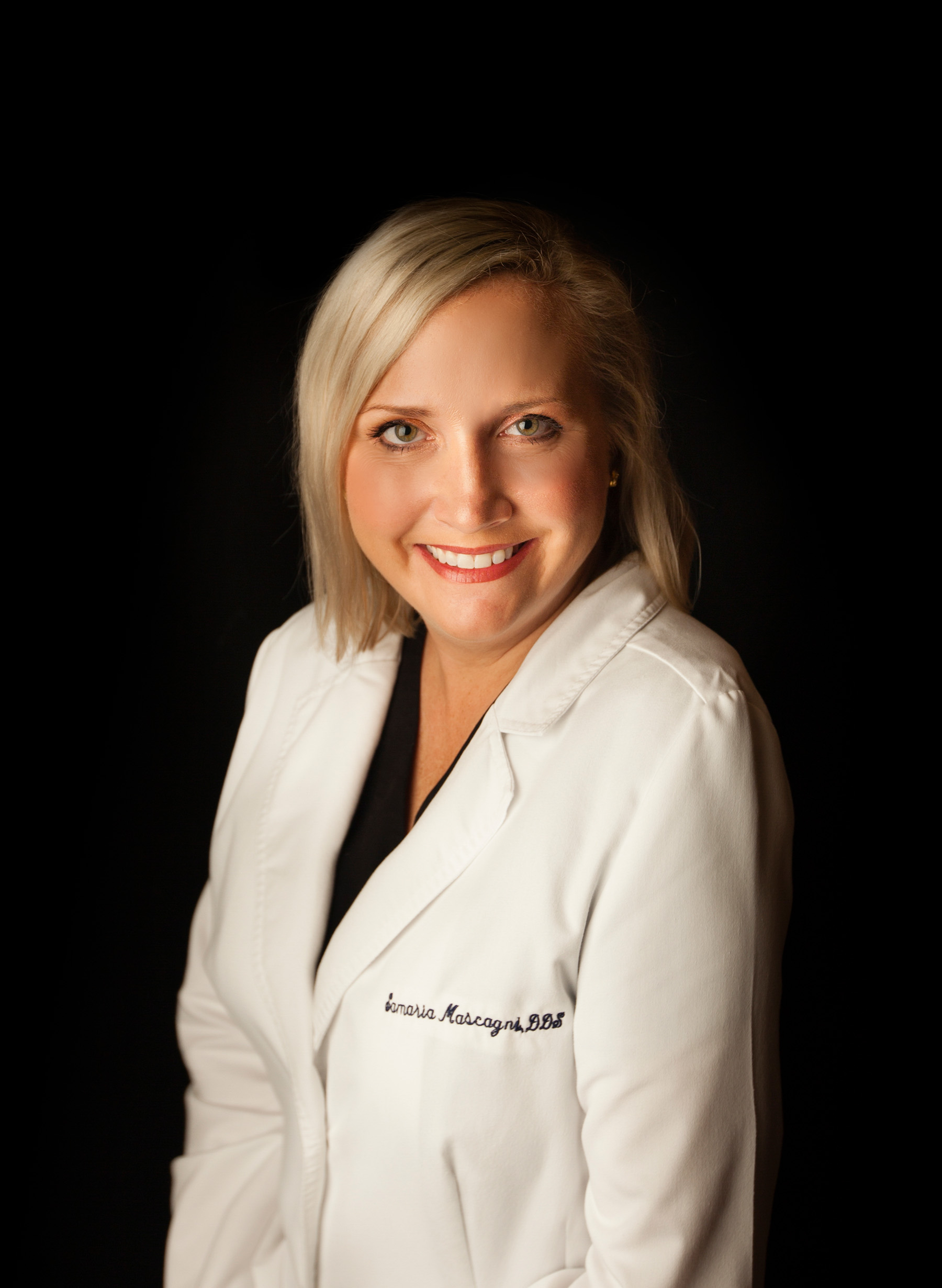 Smiling woman with blonde hair wearing a white lab coat against a black background.