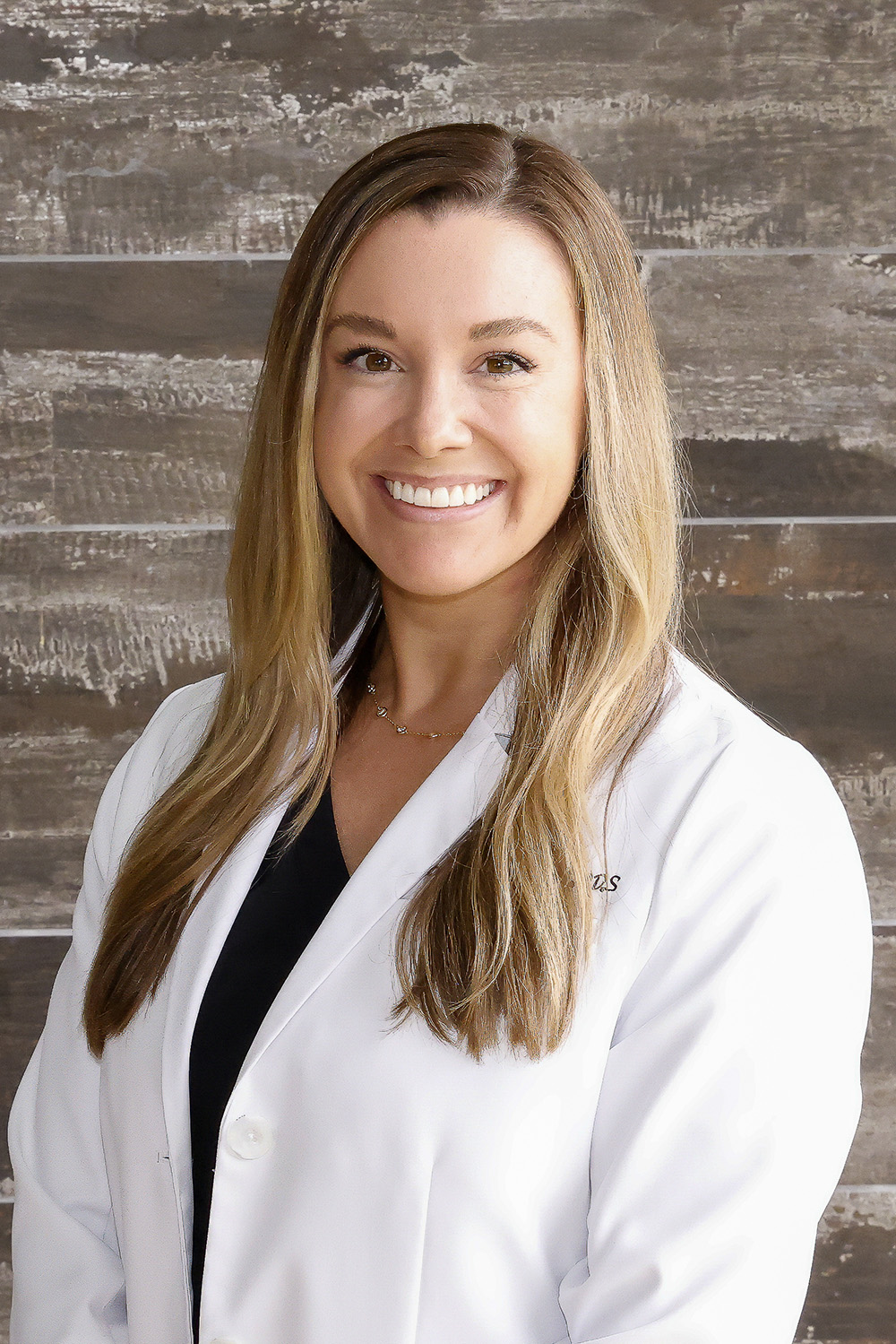 Smiling woman with long blonde hair wearing a white medical coat and black shirt, standing in front of a textured brown background.
