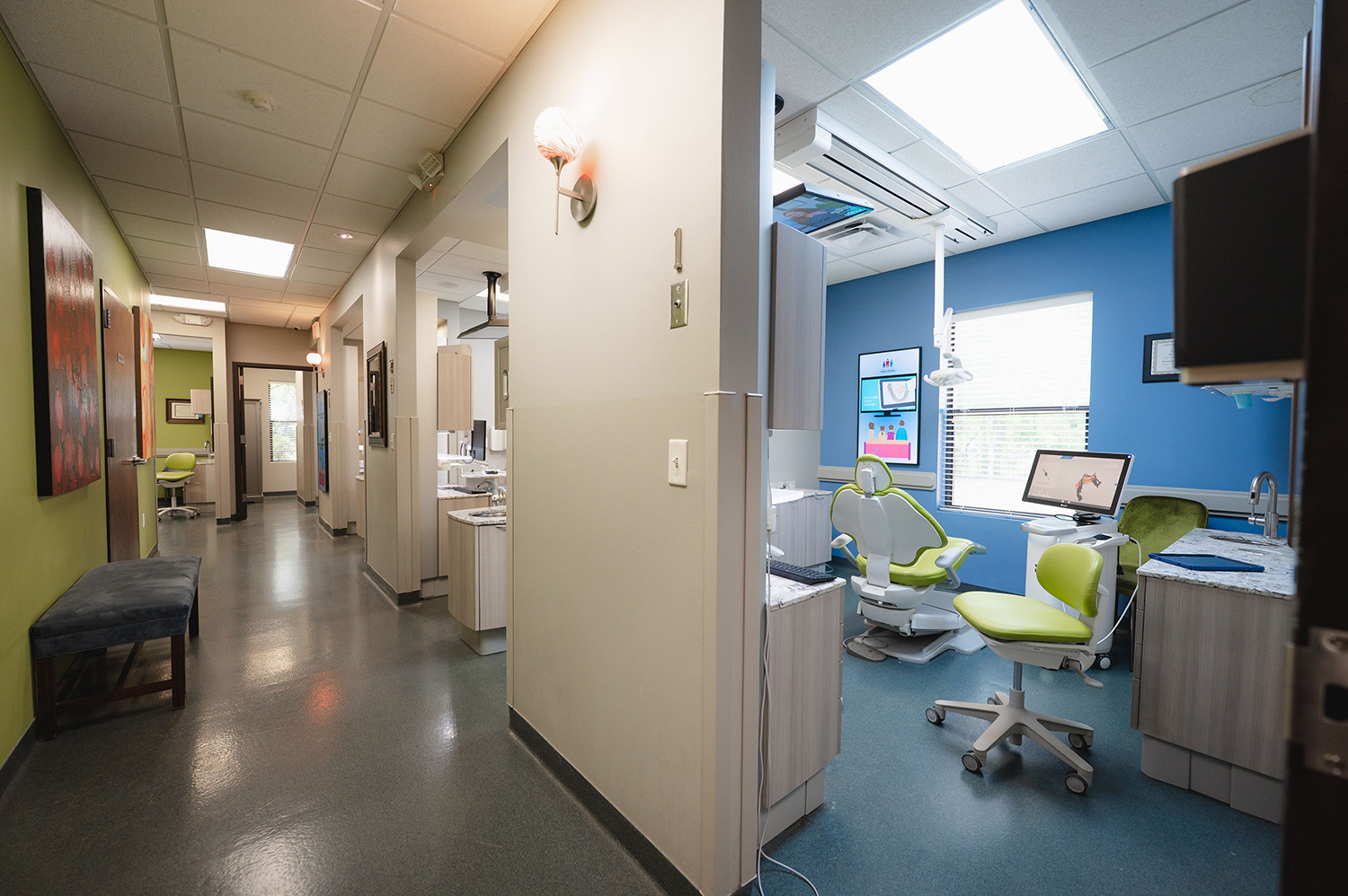 Modern dental clinic hallway leading to treatment rooms with green and blue accents and dental equipment.