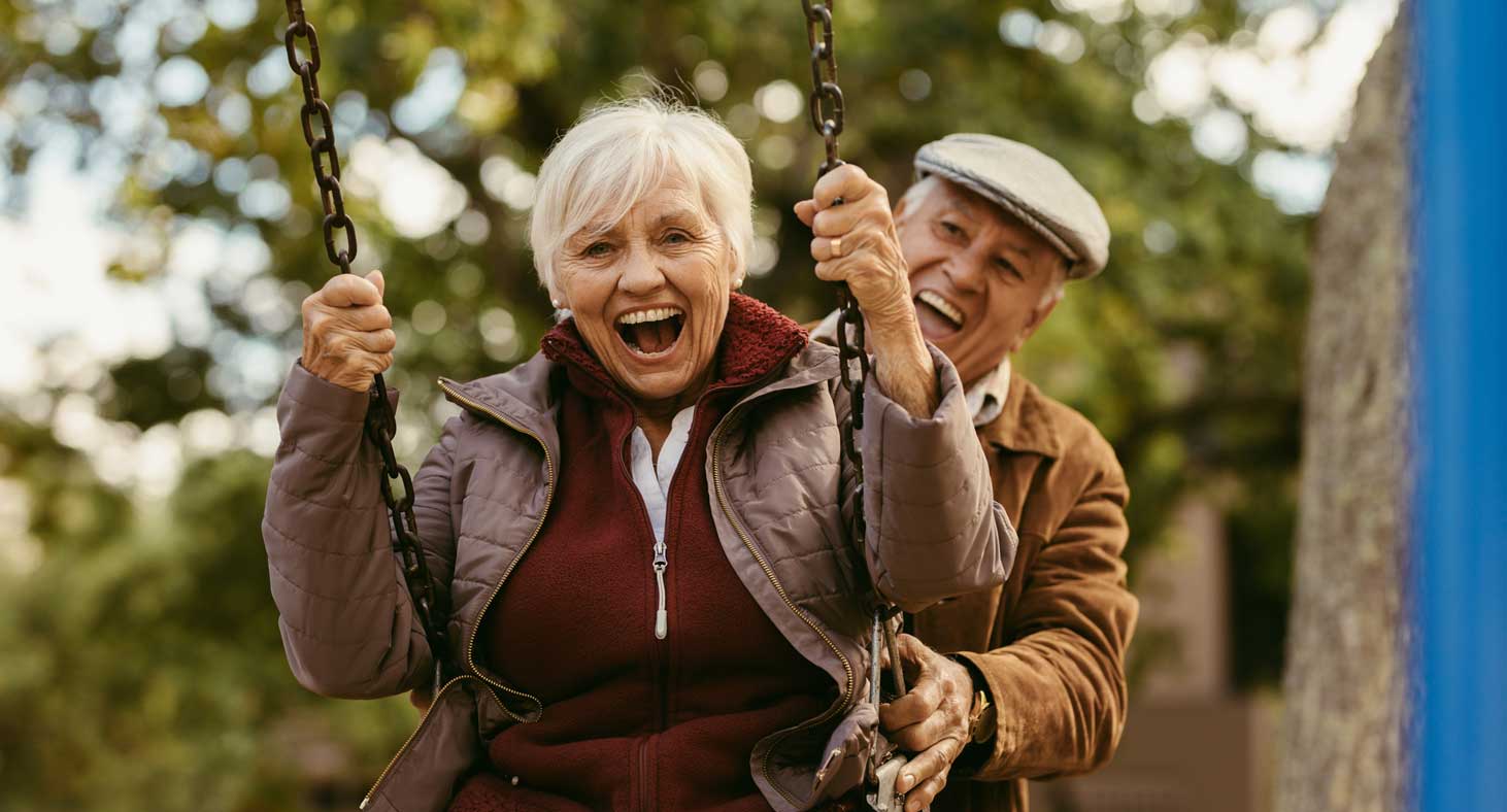 An older couple play on a swing, smile broadly to show off their mini dental implants.