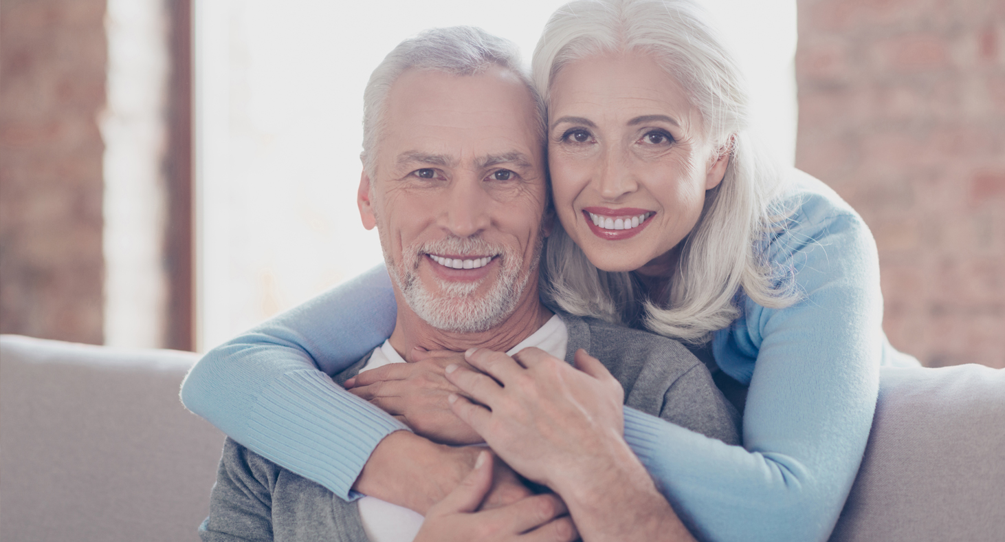 A woman leans over her husbands shoulder as they both smile, making it impossible to tell who is wearing mini dental implants.