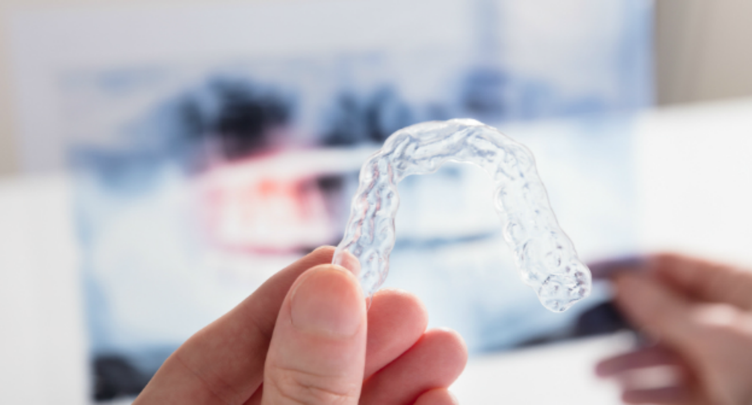 A dentist holds a clear invisalign mouth piece over a blurred x-ray.