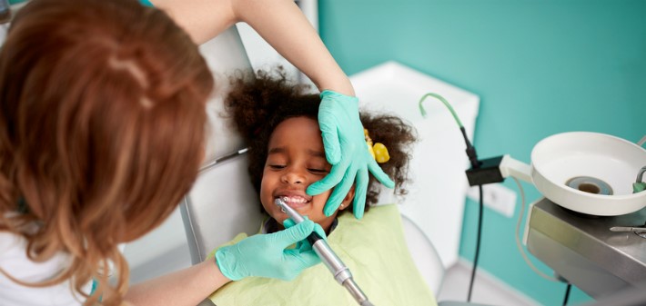 A little girl lays on the dental chair, smiling with her eyes close while her teeth are cleaned.