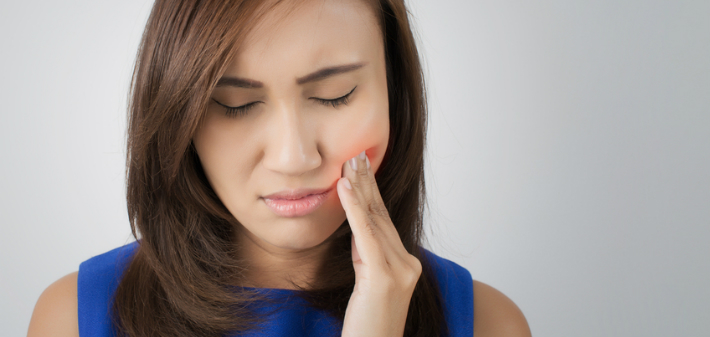 A young woman winces and rubs her cheek where her tooth aches and the surrounding gums are sore.
