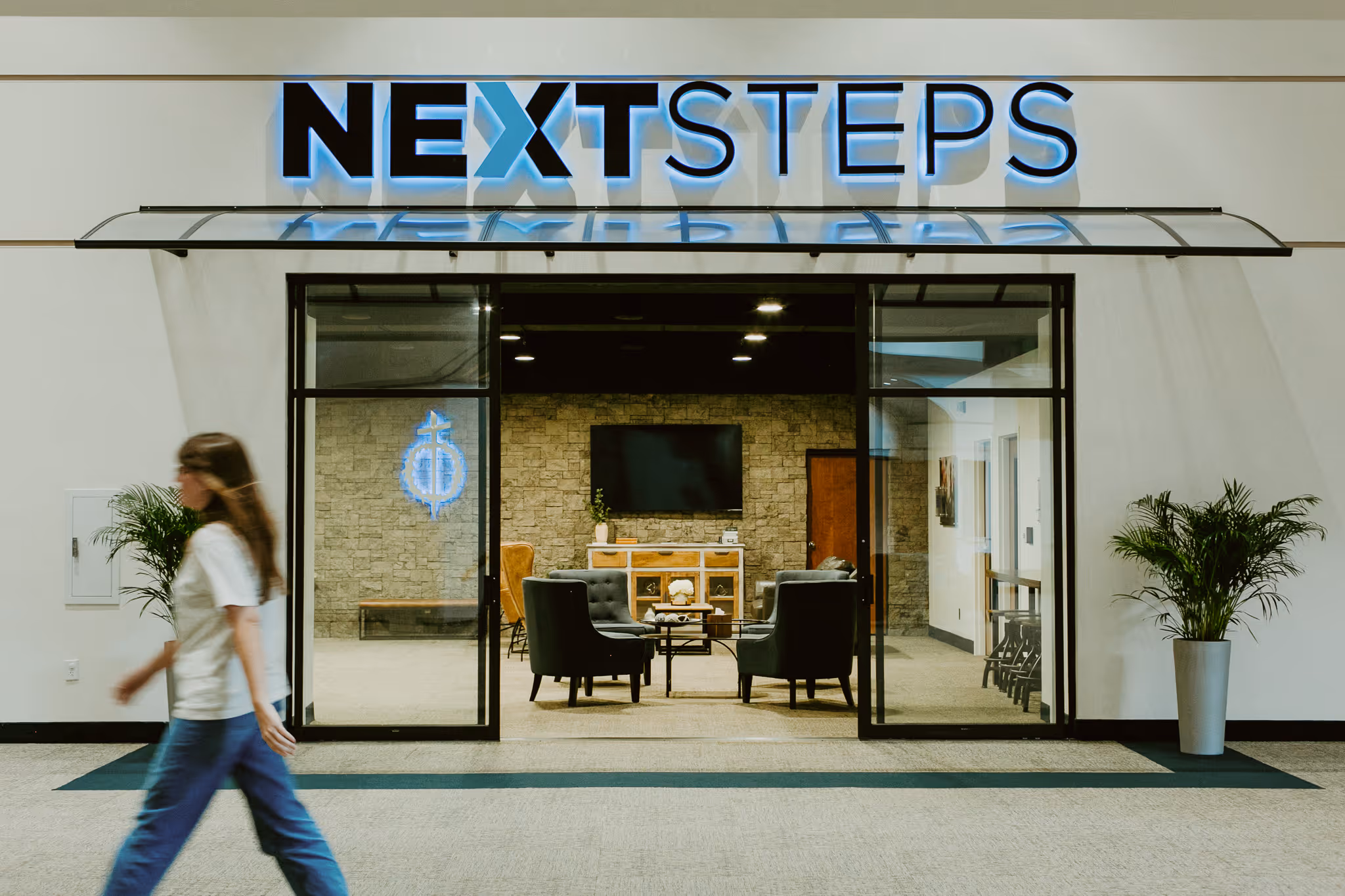 Office entrance with glass doors and illuminated NEXT STEPS sign above, featuring a seating area inside and a person walking by outside.
