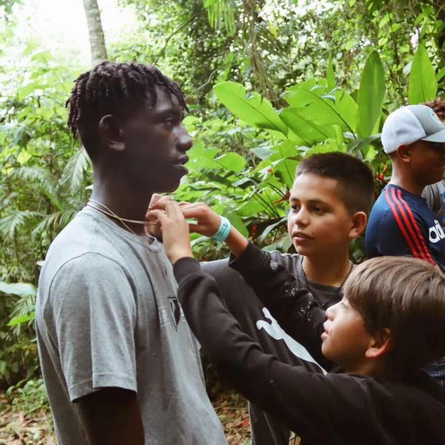 Two boys helping another boy put on a necklace in a lush, green outdoor setting.