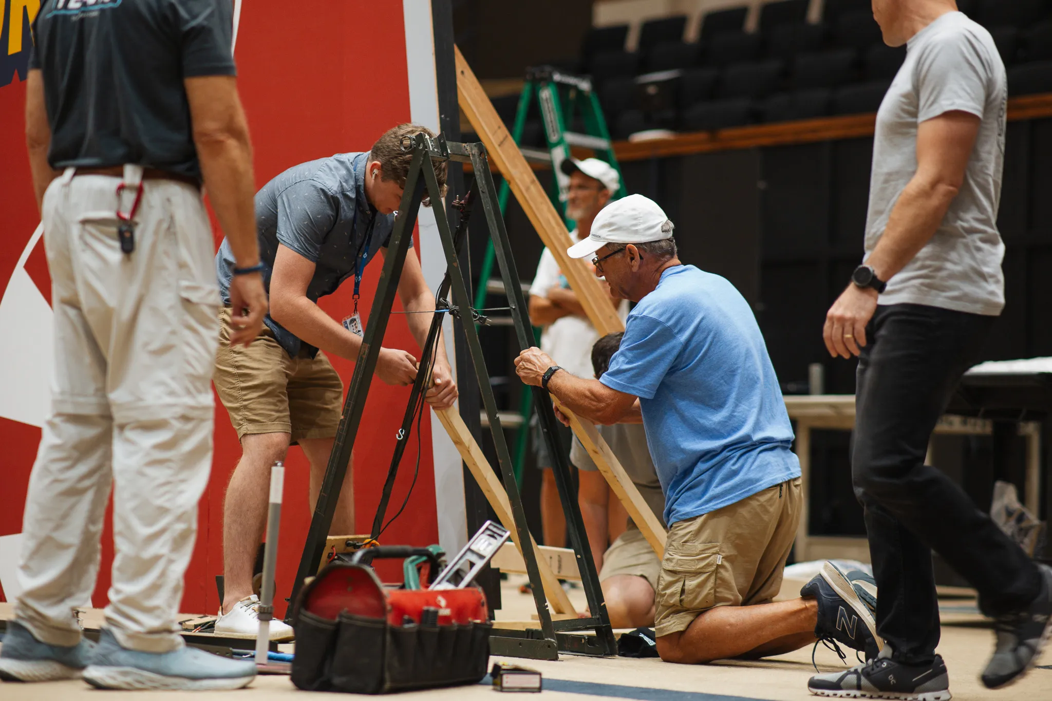Group of men assembling a wooden and metal structure indoors with tools and ladders around.