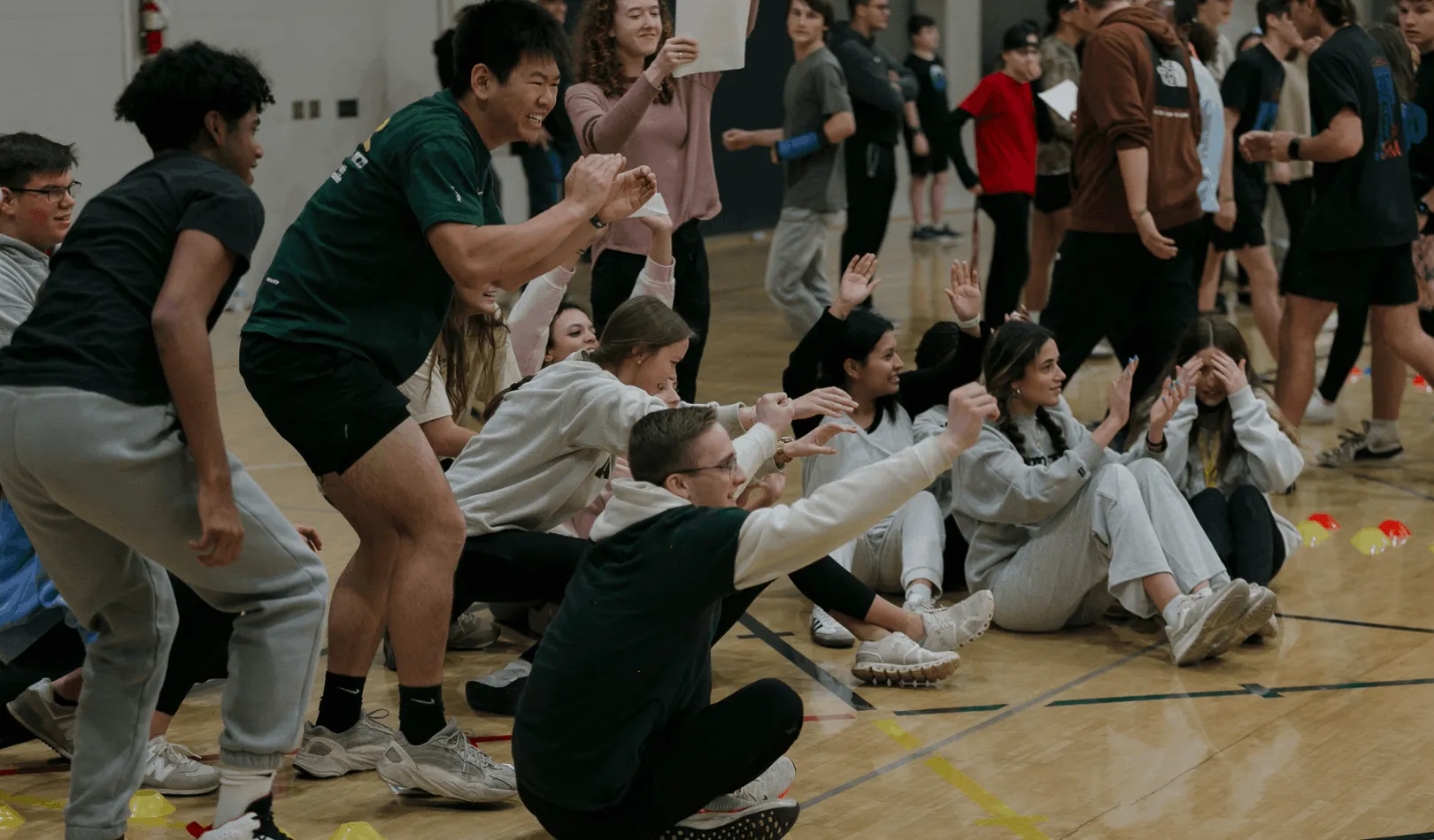 Group of diverse young people smiling and raising their hands while sitting and squatting on a gymnasium floor.