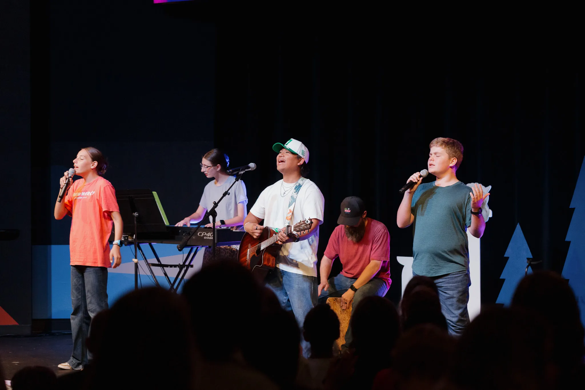 Five people on stage performing music with two singing into microphones, one playing keyboard, one playing guitar, and one playing cajón.
